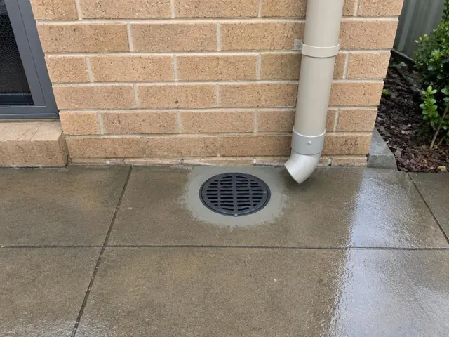 Clear outdoor drain grate beside wet paving and a downpipe against a brick wall in Marsden Park